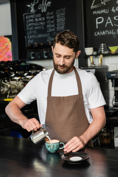 bearded barista in apron pouring milk from pitcher while making cappuccino on bar in coffee shop - Foto, Imagem