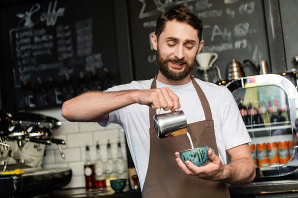 positive barista in apron pouring milk from pitcher in cup with cappuccino in coffee shop - Photo, Image