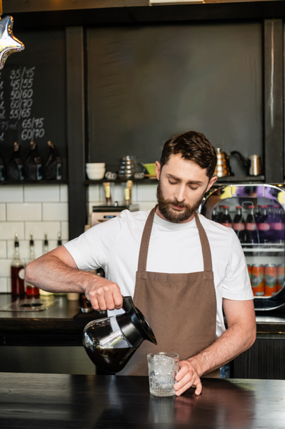 barista en delantal verter el café de la olla en el vaso con cubitos de hielo en el bar en la cafetería - Foto, Imagen