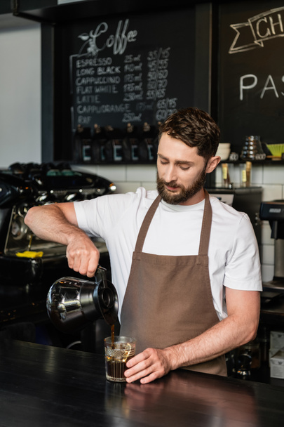 γενειοφόρος barista ρίχνει καφέ από την κατσαρόλα σε ποτήρι με παγάκια, μπαρ σε καφετέρια, καφές πάγο - Φωτογραφία, εικόνα