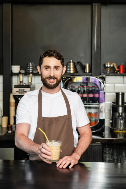 smiling barista in apron holding cold coffee drink with straw and looking at camera in coffee shop - Photo, Image