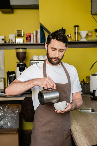 barista concentrato in grembiule versando latte in tazza con caffè mentre lavorava in caffetteria - Foto, immagini