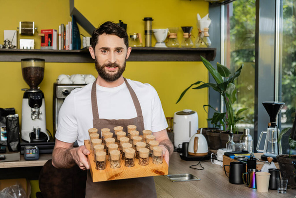 smiling barista in apron holding jars with coffee beans and looking at camera in coffee shop - Photo, Image