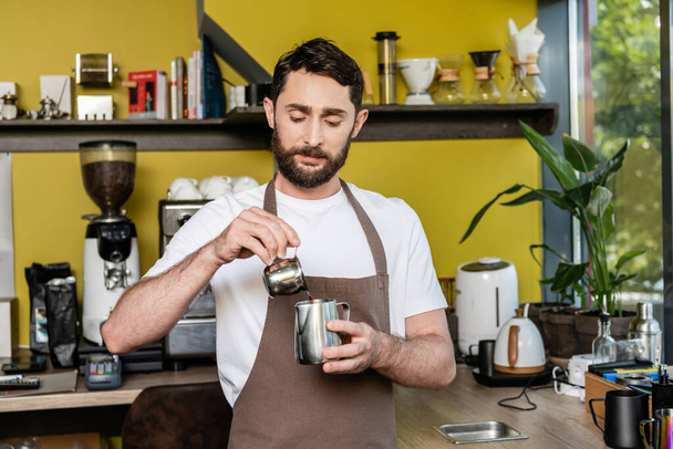 barbudo barista en delantal verter café en jarra de metal mientras se trabaja en la cafetería en el fondo - Foto, imagen