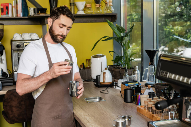 Kafede kahve yaparken elinde metal sürahi tutan önlüklü sakallı barista. - Fotoğraf, Görsel