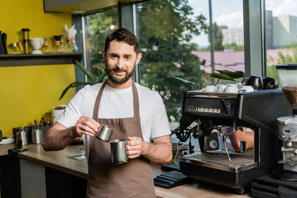 cheerful bearded barista in apron holding pitchers near coffee machine in coffee shop - Photo, Image