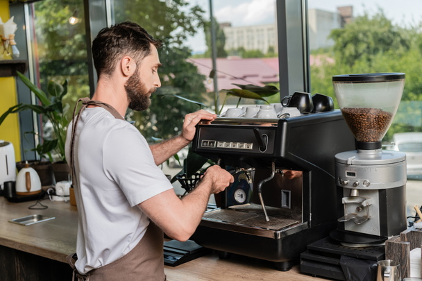 вид сбоку бородатой баристы в фартуке, работающей с кофемашиной в кафе - Фото, изображение