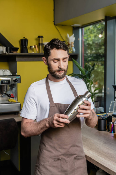 szakállas barista kötényben tartja shaker miközben áll kávéfőző mellett kávézóban - Fotó, kép