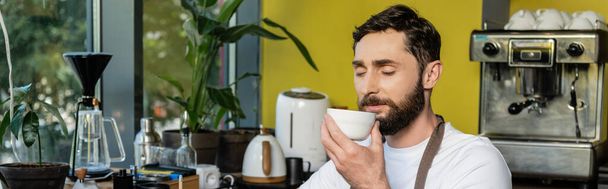 бородатый бариста в фартуке нюхает кофе в чашке во время работы в кафе, баннер - Фото, изображение