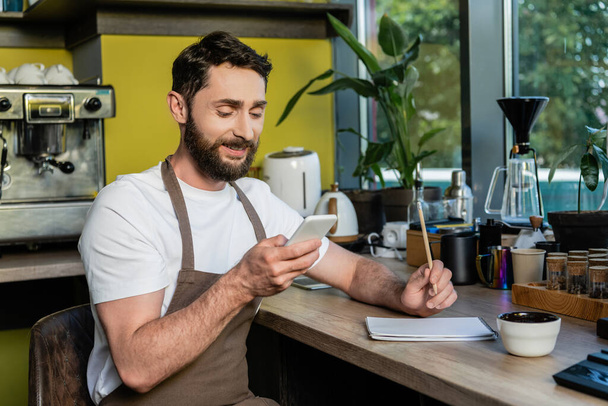 positiivinen barista esiliina älypuhelimella lähellä kannettavaa työtasoa kahvilassa - Valokuva, kuva