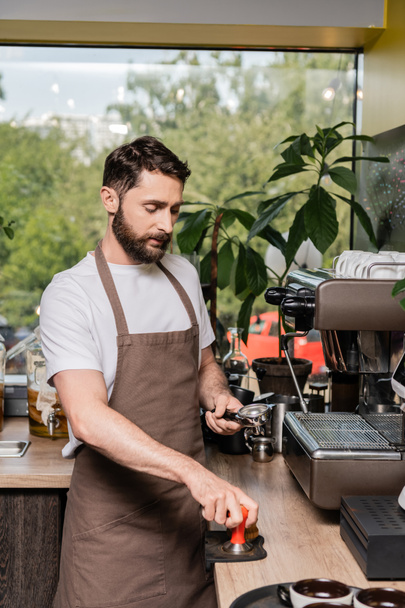 barista met baard in schort met pers en houder in de buurt van koffiezetapparaat in coffeeshop - Foto, afbeelding