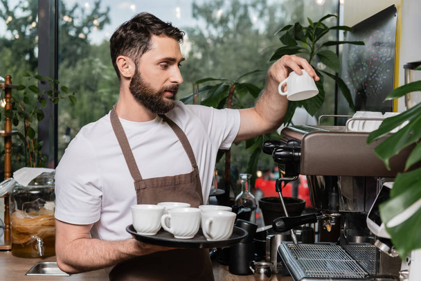 szakállas barista kötényben tiszta csészéket tesz a kávéfőzőre, miközben a kávézóban dolgozik. - Fotó, kép