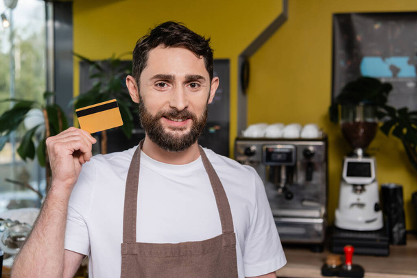 mosolygós barista kötényben nézi a kamerát, és tartja hitelkártya kávézóban - Fotó, kép