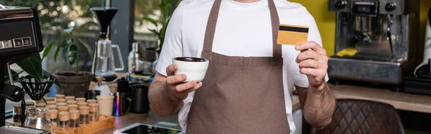 vista cortada de barista em avental segurando cartão de crédito e xícara enquanto trabalhava em café, banner - Foto, Imagem