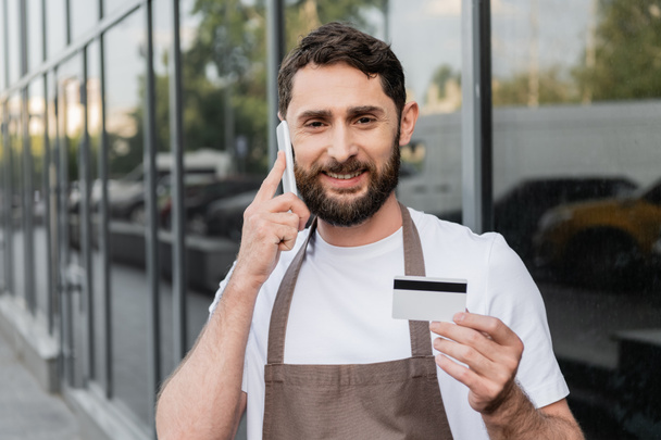 veselý barista drží kreditní kartu a mluvit na smartphone na městské ulici na pozadí - Fotografie, Obrázek