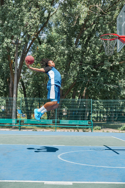 Mavi üniformalı genç adamın tam boy dinamik görüntüsü atlıyor, güneşli bir günde açık havada basketbol oynuyor. Profesyonel spor, rekabet, hobi, oyun, aktif yaşam tarzı kavramı - Fotoğraf, Görsel