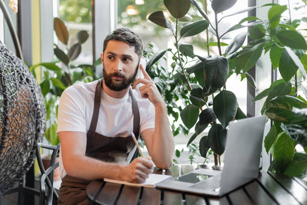 barista puhuu älypuhelimella ja kirjoittaa kannettavan lähellä kannettavaa tietokonetta ja kasveja kahvilassa - Valokuva, kuva