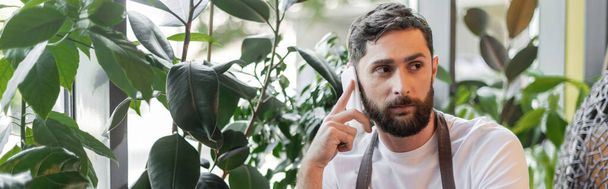 бородатий бариста в фартусі розмовляє на смартфоні, стоячи біля рослин в кав'ярні, банер - Фото, зображення