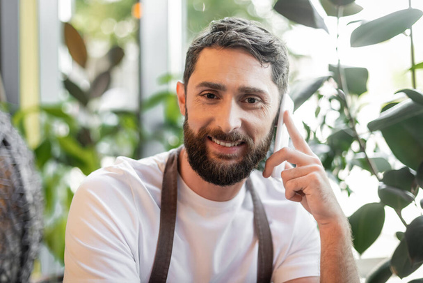 portrét pozitivního vousatého baristy mluvícího na smartphonu a dívajícího se do kamery v kavárně - Fotografie, Obrázek