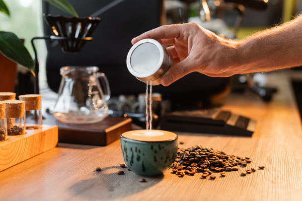 zugeschnittene Ansicht des Barista, der Zucker in eine Tasse Cappuccino in der Nähe von Kaffeebohnen gießt, im Licht des Cafés - Foto, Bild