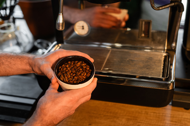cropped view of barista holding cup with coffee beans near blurred coffee machine in cafe - Photo, Image