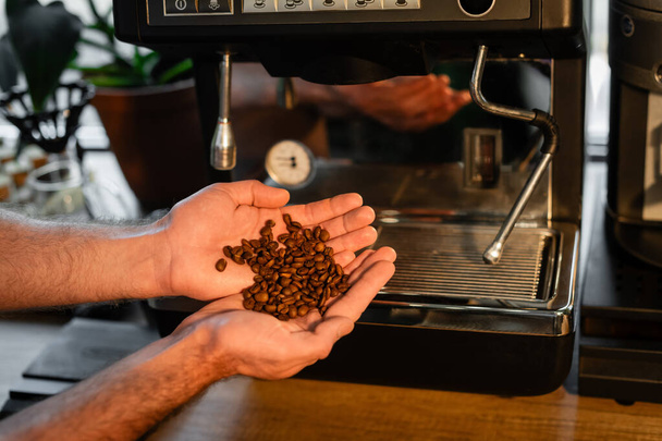 обрезанный вид на бариста с кофейными зернами на руках возле кофеварки в кафе - Фото, изображение