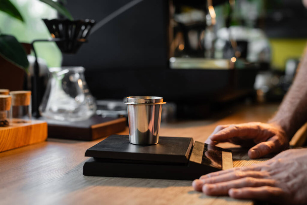 καλλιεργημένη άποψη του barista στέκεται κοντά σε ποτήρι ζέσεως σε ηλεκτρονική κλίμακα κοντά σε καφετιέρα στο καφέ - Φωτογραφία, εικόνα