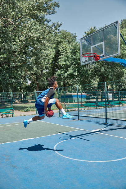 Slam dunk. Full-length image of young man in motion, playing basketball outdoors on sportsground on warm day. Concept of professional sport, competition, hobby, game, active lifestyle - Foto, afbeelding