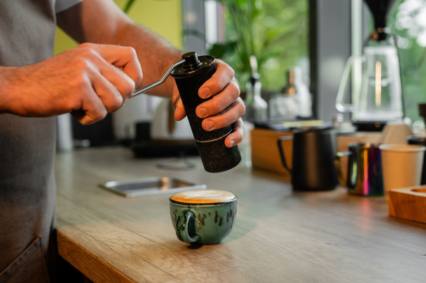 обрезанный вид на шлифовальную корицу бариста над чашкой капучино на столешнице в кафе - Фото, изображение