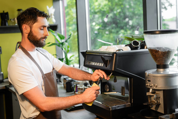 Bärtiger Barista reinigt Kaffeemaschinen-Düse mit Lappen bei der Arbeit im Café - Foto, Bild