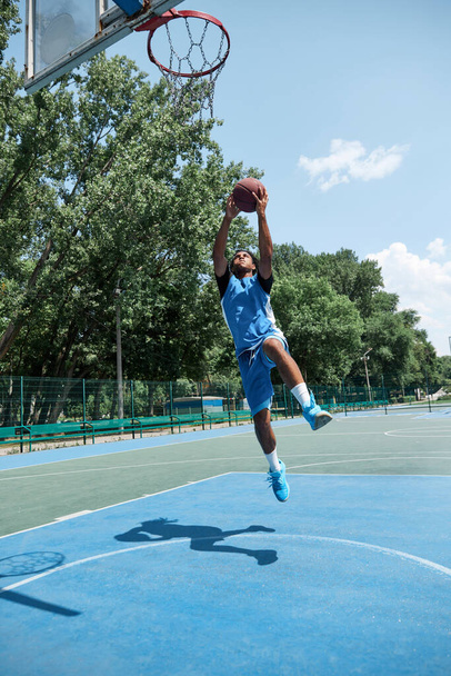 Full-length dynamic image of young guy in blue uniform jumping, playing basketball outdoors on sportsground on warm sunny day. Concept of professional sport, competition, hobby, game, active lifestyle - Fotoğraf, Görsel