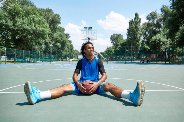 Young guy in blue uniform, basketball player sitting and resting after game. Outdoor sportsground. Concept of professional sport, competition, hobby, game, active lifestyle - 写真・画像
