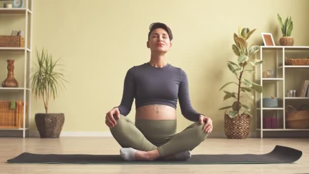 Slowmo of beautiful young pregnant woman with short dark hair and tattoo on her belly practicing yoga at home, smiling at camera - Imágenes, Vídeo