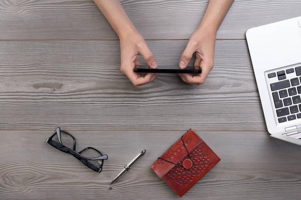 Top view of a table surface, young unidentifiable person's hands using a smartphone for work or leisure. Notebook, pen, glasses, and a laptop present - Photo, Image