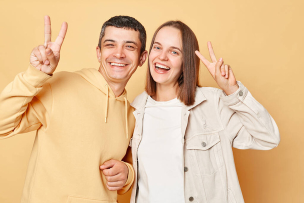 Happy cheerful woman and man wearing casual style clothing standing isolated over beige background looking smiling to camera, showing v signs, victory gesture. - Fotoğraf, Görsel