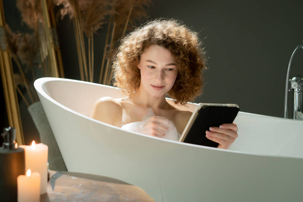 魅力的な若い女性がお風呂に横たわり,デジタルタブレットで何かを見ています. 美しい女性は夜の風呂に入っている間,彼女のタブレットにソーシャルメディアを閲覧しています. 高品質の写真 - 写真・画像
