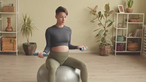 Energetic young pregnant woman exercising with dumbbells while sitting on fit ball having indoor fitness training - Video