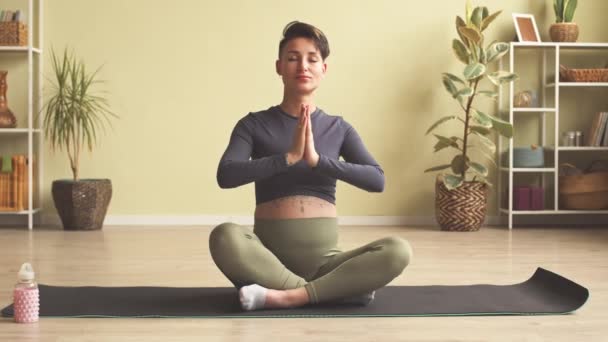 Slowmo of modern young pregnant woman with short haircut and tattoo on her belly meditating on yoga mat at home - Imágenes, Vídeo