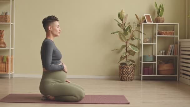 Slowmo of concentrated young pregnant woman sitting on her laps on yoga mat touching her belly and practicing breathing - Felvétel, videó