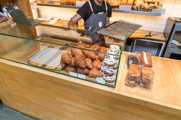 Bustling bakery scene with a worker replenishing croissants using tongs, surrounded by diverse bread shelves and sweet treats. - 写真・画像