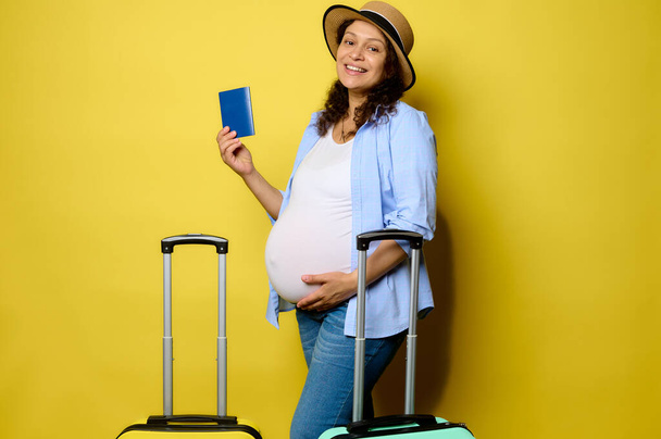 Multi-ethnic gorgeous smiling charming pregnant woman traveler passenger in casual clothes, puts her hand on her belly, shows a passport document, looks at camera, isolated on yellow studio background - Foto, immagini