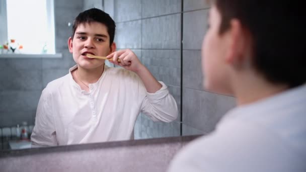 Charmanter Teenager kümmert sich um Mundhygiene und putzt sich im Badezimmer die Zähne mit Zahnpasta und Bürste, schaut in den Spiegel - Filmmaterial, Video