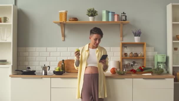 Medium shot of young smiling pregnant woman in relaxed light yellow shirt eating apple and scrolling on smartphone standing at cozy bright kitchen at leisure time - Πλάνα, βίντεο