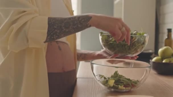 Cropped shot of young expectant mother with tattoos on her body making green salad at kitchen, adding arugula leaves into glass bowl - Filmagem, Vídeo