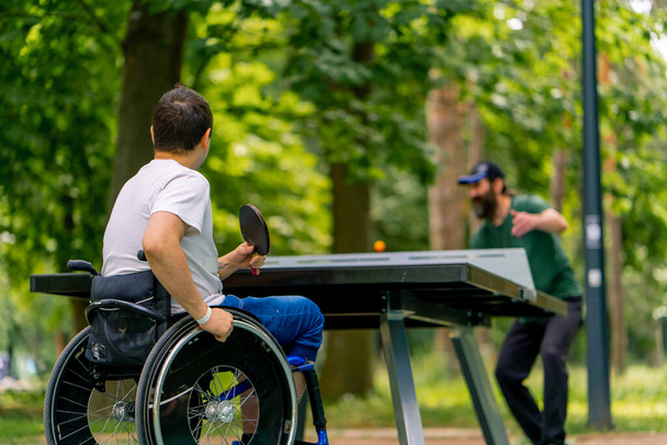 Инклюзивность Инвалид в инвалидной коляске играет в пинг-понг со взрослым мужчиной в городском парке на фоне деревьев - Фото, изображение