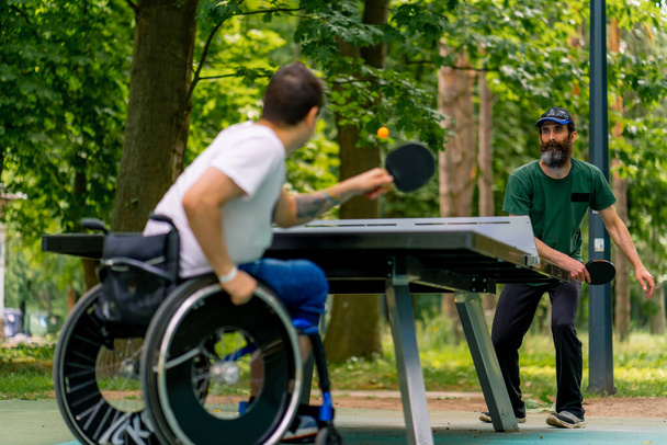 Osallisuus Vammainen mies pyörätuolissa pelaa pingistä vanhemman miehen kanssa puistossa puiden taustaa vasten - Valokuva, kuva