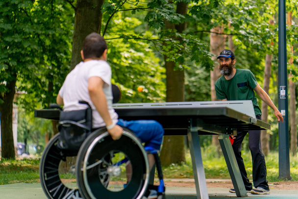 Inkluzivita Osoba se zdravotním postižením na invalidním vozíku hraje ping pong se starším mužem v městském parku na pozadí stromů - Fotografie, Obrázek