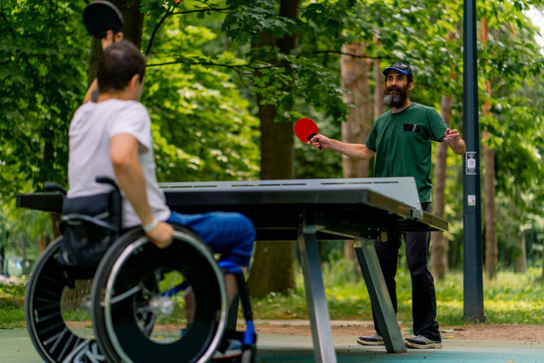 Integracja Niepełnosprawny na wózku inwalidzkim gra w ping ponga ze starszym mężczyzną w parku miejskim na tle drzew - Zdjęcie, obraz