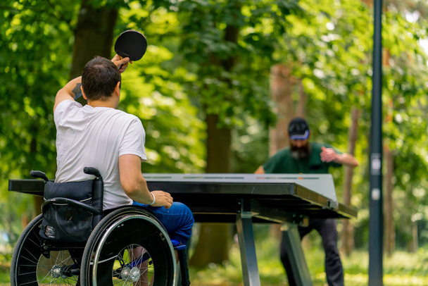 Inklusion Ein behinderter Mann im Rollstuhl spielt im Stadtpark vor Baumkulisse mit einem älteren Mann Tischtennis - Foto, Bild