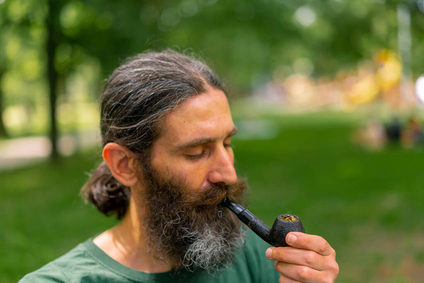 Porträt eines alten Mannes mit langen Haaren und grauem Bart, der im Stadtpark vor dem Hintergrund von Bäumen eine Pfeife raucht - Foto, Bild